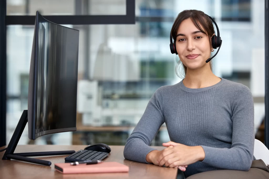 Fotografía de una mujer de suéter gris utilizando audífonos y micrófono sentada en mobiliario para call center.