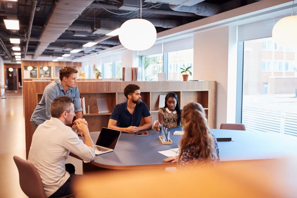 Fotografía de 4 profesionales sentados en un escritorio conversando en un espacio de coworking a lado de una ventana.